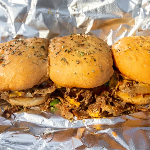 Three Tastee Buddies cheesesteak sliders with seasoned buns, steak, cheese, and onions in foil.