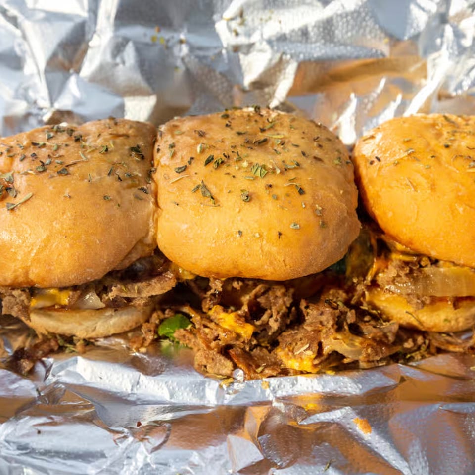 Three Tastee Buddies cheesesteak sliders with seasoned buns, steak, cheese, and onions in foil.