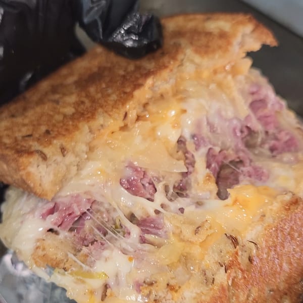 Close-up of a grilled sandwich with melted cheese, corned beef, and sauerkraut, held by a gloved hand.